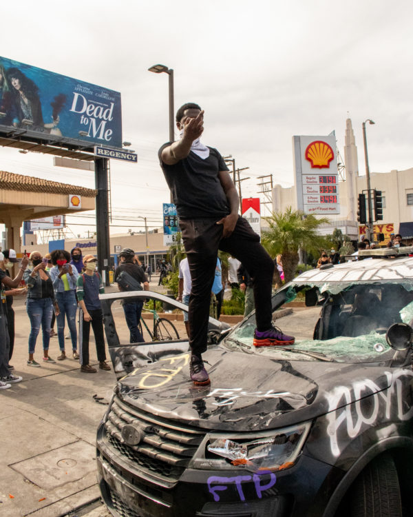 Raw and unapologetic, this image captures the visceral defiance of a man standing on top of a shattered symbol of state power. The graffiti, the cracked windshield, the unbothered stare—it’s not vandalism, it’s protest in its purest form: angry, human, and unfiltered.