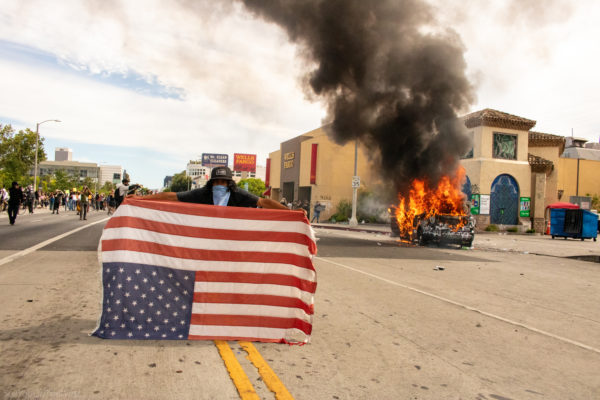 An iconic image of resistance. A protester stands firm in the middle of chaos, holding an upside-down American flag—an internationally recognized signal of dire distress. Behind him, a police vehicle burns, symbolizing the fury of generations ignored. This moment captures the duality of patriotism and pain.